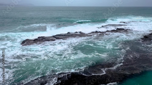 A Wide shot of ocean waves crashing over black rocky coastline with turquoise foam