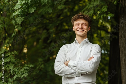 Smiling young man with curly hair stands with his arms crossed outdoors in a leafy green forest setting.