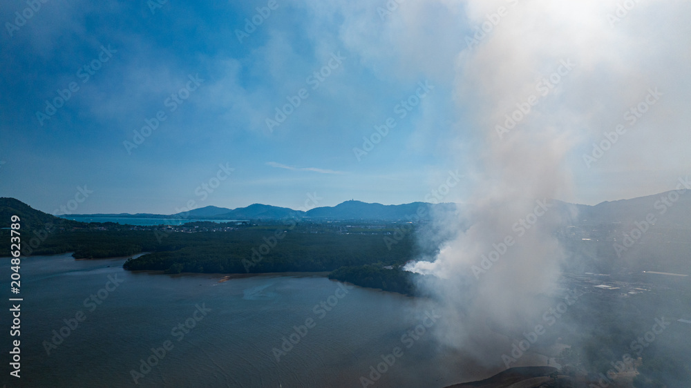 Obraz premium Aerial view of a landfill fire in Phuket with heavy smoke spreading across nearby landscapes and urban areas. Thick plumes of smoke rise illustrating the impact of urban waste and air pollution.