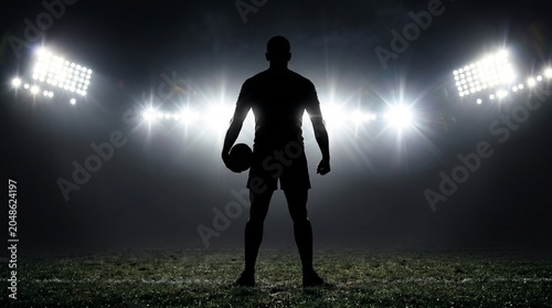 Silhouette of a determined rugby player standing on a stadium pitch under bright spotlights at night