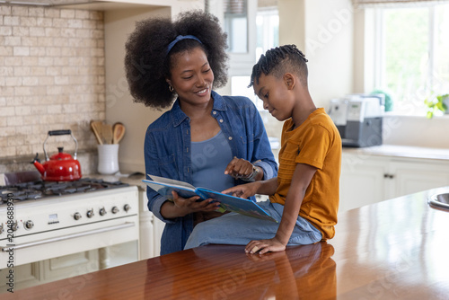 African American mother in denim shirt and son reading picture book on wooden countertop in kitchen