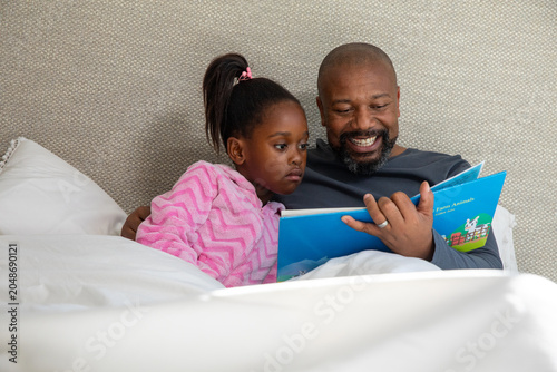 African American father and daughter in pink pajamas reading blue book on white bedding, smiling
