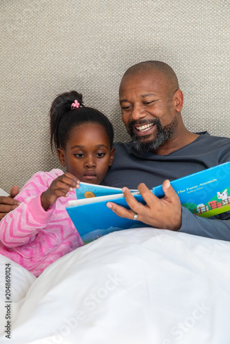 African American father and child reading Farm Animals book on bed, child wearing pink pajamas