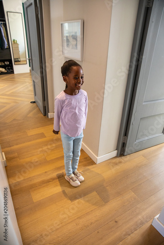 African American child girl smiling in hallway wearing light top and sneakers near framed artwork