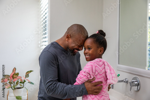 African American father and child pressing foreheads at bathroom sink in pink robe and gray shirt