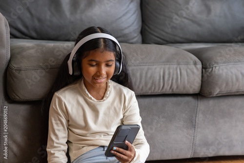 Indian child girl sitting on gray sofa at home, holding smartphone wearing over-ear headphones