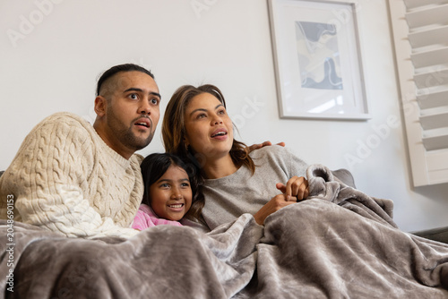 Family parents with child sitting on couch at home under gray blanket wearing sweaters pink top