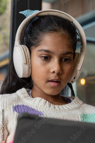 Indian child wearing beige headphones and cream knit sweater, looking down at tablet near window