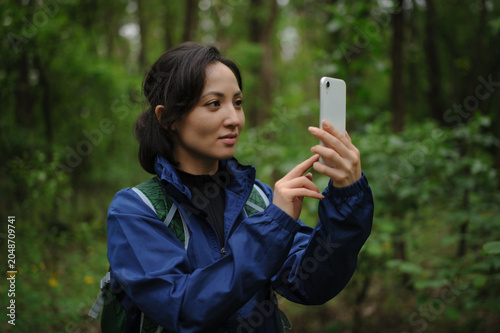 Portrait of asian tourist with backpack taking photo on smartphone in forest 
