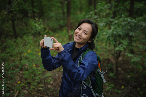 Smiling asian woman backpacker taking photo on smartphone and looking at camera in forest 