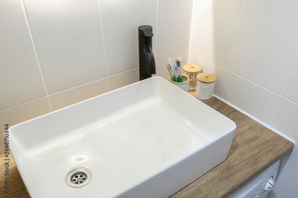 Fototapeta premium white rectangular vessel sink sits atop wooden countertop; paired with matte black faucet, against white tiled walls, with bamboo containers holding toiletries nearby