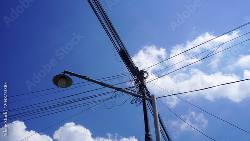 Tangled electric cables and street lamp pole against bright blue sky with clouds.