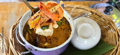Traditional clear tom yum soup with shrimp is served in a small purple tin pot with separate bowls, placed on a brown woven basket. Use a spoon to scoop out the large shrimp, King Trumpet Mushroom.
