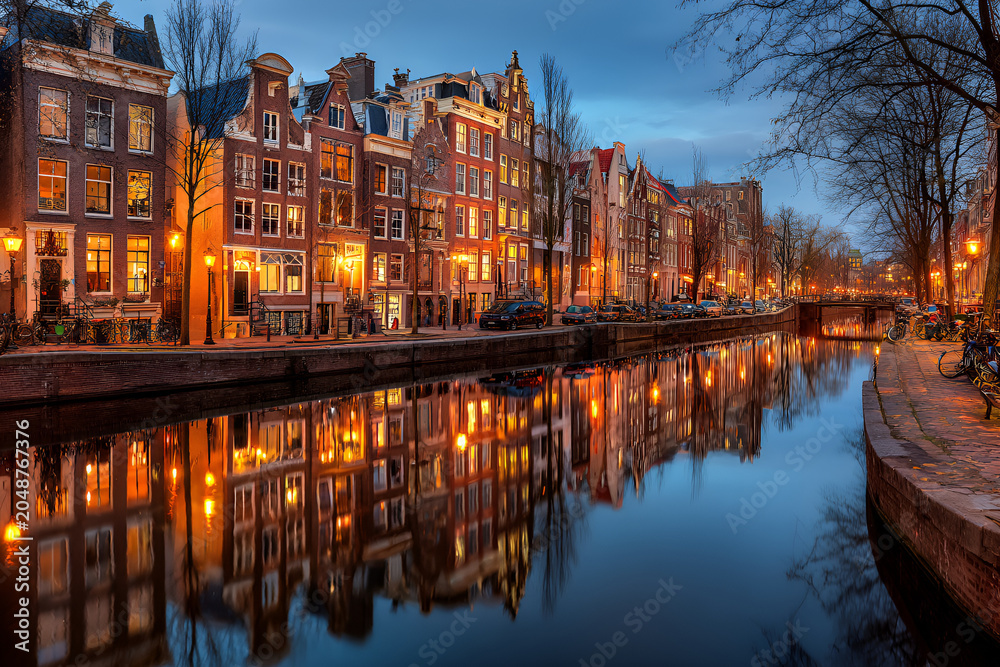 Fototapeta premium Illuminated historical buildings line a tranquil canal, reflecting lights in the water at twilight. Trees frame the scene