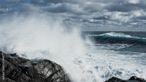 Powerful ocean waves crashing against jagged black coastal rocks under a cloudy dramatic sky