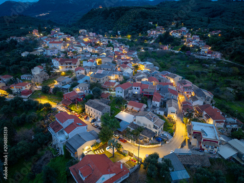 Aerial view of illuminated Nimfes village at dusk, featuring traditional architecture and winding roads in the mountains of Corfu island, Greece