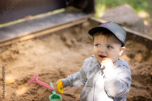 Obraz na plátně Young child playing in sandbox wearing gray jacket and black cap, holding a toy