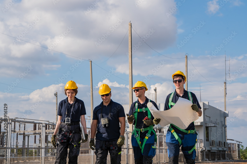 Fototapeta premium Four workers in hardhats and harnesses walk through a power substation. They hold a blueprint and discuss infrastructure plans. The diverse team focuses on renewable energy industry growth