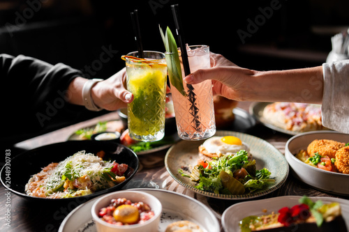 Quadro su tela Two people clink glasses of colorful cocktails over a restaurant table filled with delicious dishes like salads, tartare, and main courses