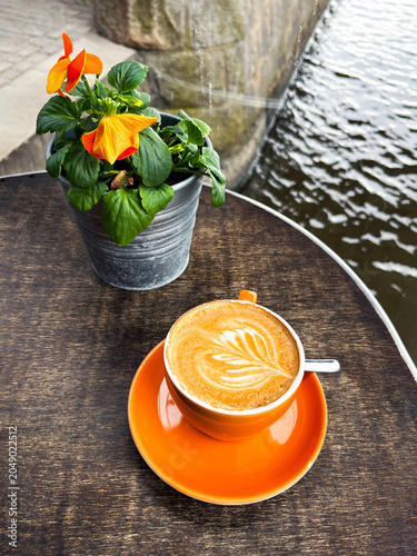 Cup of Hot Cappuccino Coffee and Flower on Table in a Cafeteria on the River Bank.