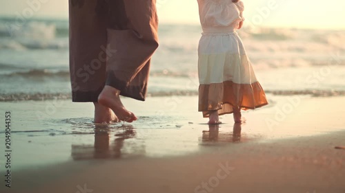 Close up of Asian parent and toddler girl feet walking barefoot on sandy beach at sunset golden hour. Hazy cinematic shot of family summer vacation and slow living bonding. Ideal for travel, parenting