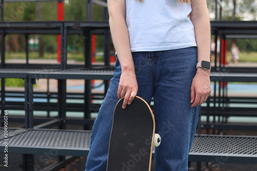 Tableau sur toile Girl in blue jeans and white t-shirt holding skateboard, standing against city tribunes background