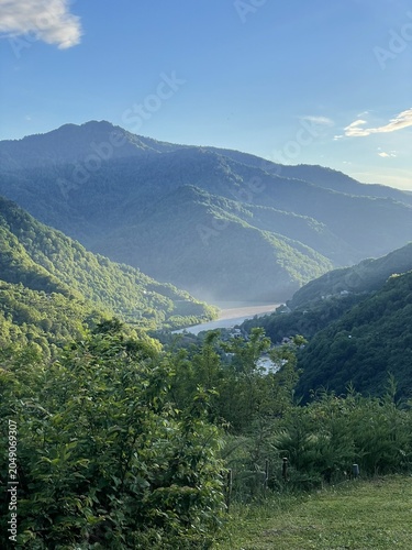 A scenic view of a lush green valley surrounded by mountains under a clear blue sky. The sunlight casts a soft glow on the landscape.