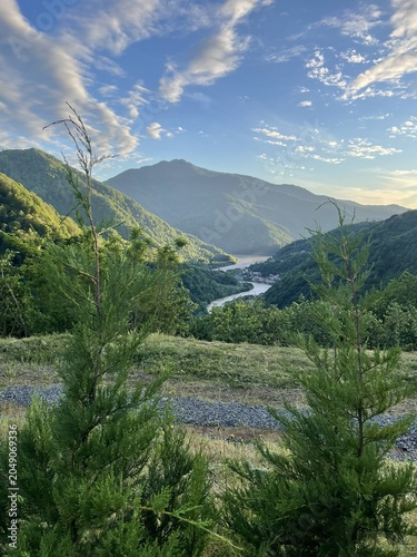A scenic view of mountains and a river surrounded by lush greenery. The sky is partly cloudy with a warm light illuminating the landscape.