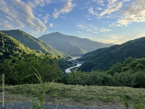 A scenic view of a river winding through lush green mountains under a blue sky with scattered clouds. The landscape showcases natural beauty and tranquility.