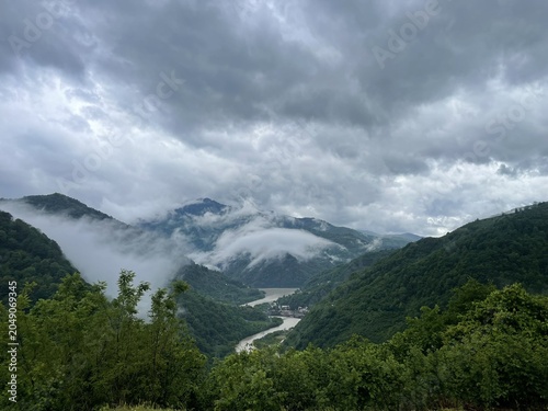 A scenic view of mountains under a cloudy sky. Lush green hills surround a winding river, with mist hovering over the landscape.