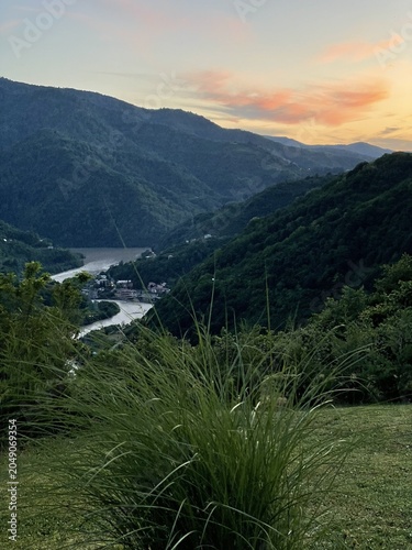 A scenic view of mountains at sunset, with a river winding through the valley. Lush green grass in the foreground adds to the natural beauty.