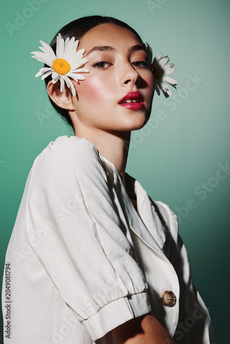 Canvastavla Young woman with floral accessories in hair wears a white jacket and shiny makeup, posing confidently against a pale green background in a creative beauty fashion studio shot