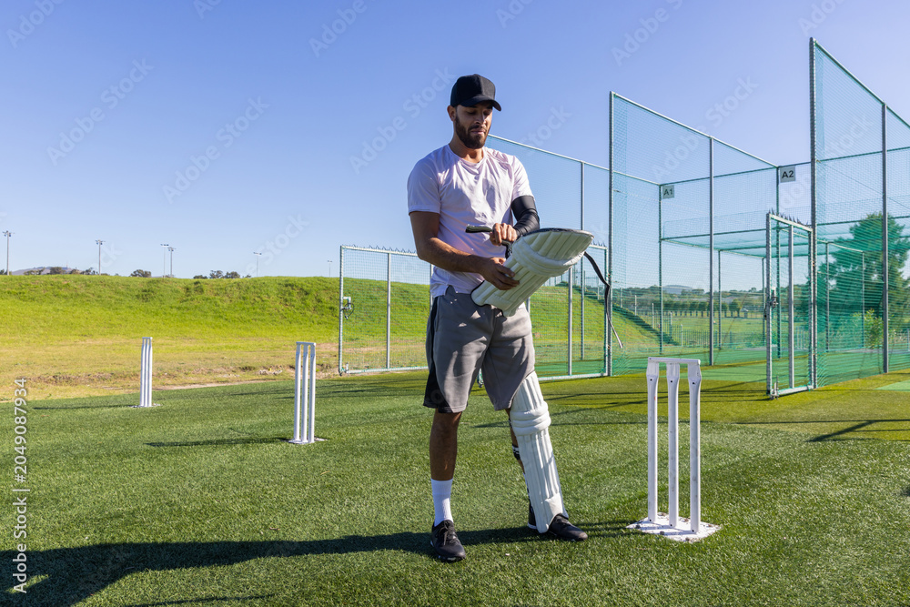 Obraz premium Man adjusting batting pad while standing on turf at cricket nets with stumps nearby