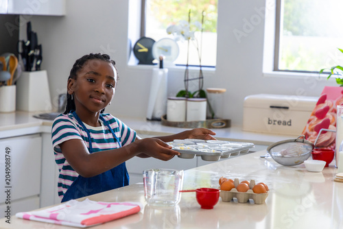 Foto Preteen African girl wearing blue apron holding muffin tray with liners in home