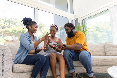 African family playing hand game on beige sofa by large windows, wearing denim and mustard shirts