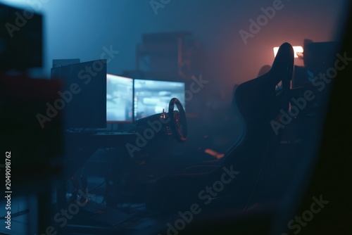 Realistic sim racing scene featuring professional gaming setup with multiple screens, racing wheel, and gaming chair in a dimly lit environment during a virtual racing competition