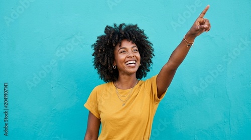 Cheerful African American Woman with Natural Curly Hair Pointing Up on Turquoise Background – Copy Space