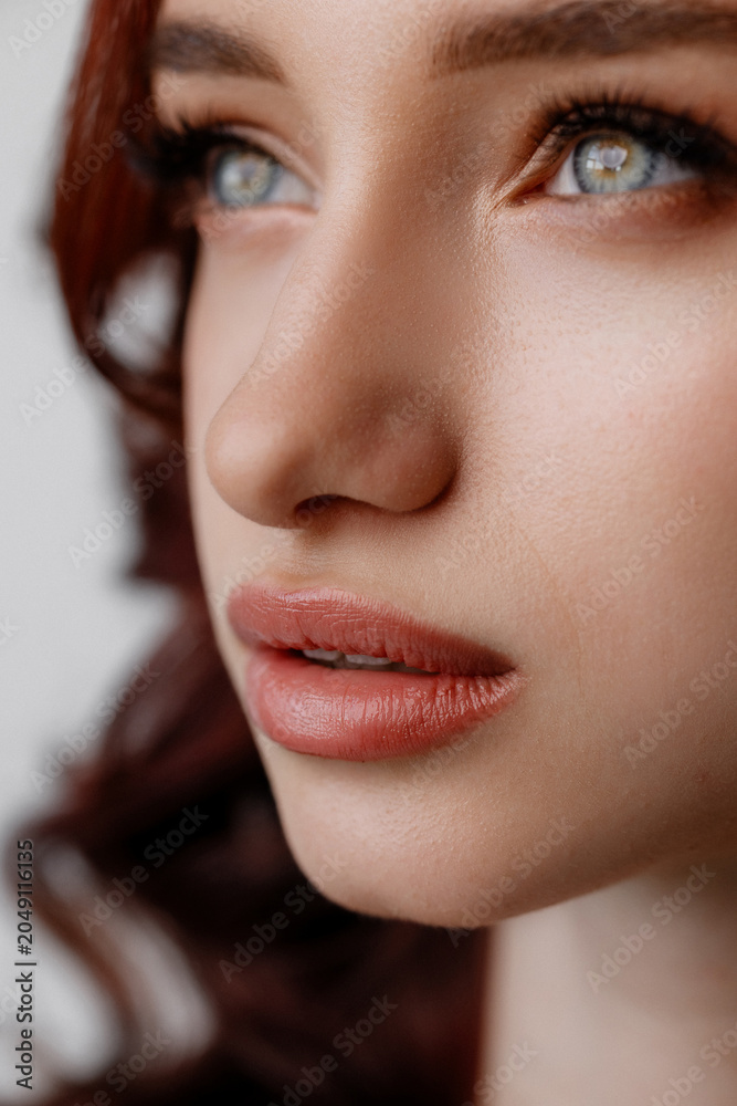 Fototapeta premium Detailed close-up of a womans face with soft natural makeup, clear blue eyes, defined lips, and smooth skin against a blurred neutral background with gentle