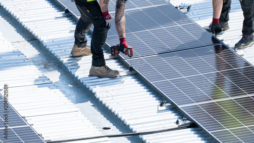 Obraz na płótnie Multiple worker using drill to fasten solar panel row on bright rooftop