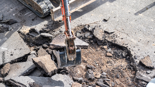 Excavator bucket digging into broken asphalt hole with rubble and gravel debris