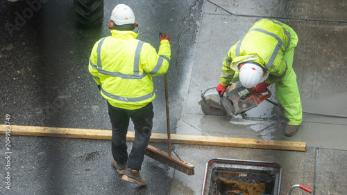 Two construction workers in yellow vest cutting concrete pavement road from overhead view