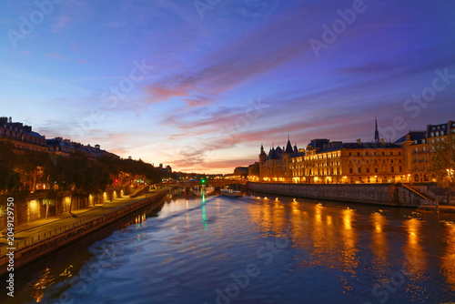 Breathtaking blue-hour panorama of the River Seine in Paris, with the illuminated Conciergerie and its medieval towers glowing in golden light on the right bank.