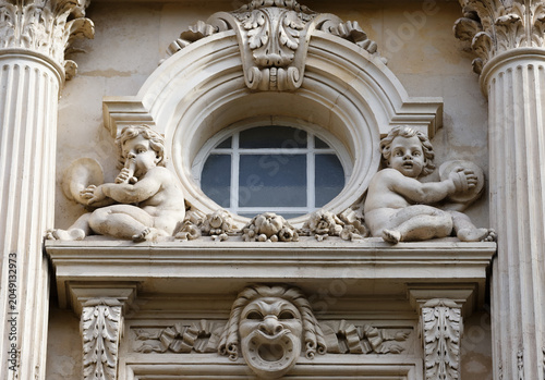 Exquisite architectural detail of a classical Parisian building facade featuring two playful limestone putti — cherubs playing musical instruments — flanking a circular bull's-eye window. 