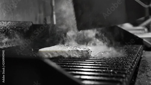 Black and white close up of a burger patty being flipped on a hot grill with steam rising