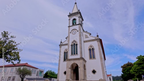 Low angle view of the traditional Catholic church facade against a blue sky with soft wispy clouds. The white building features stone trimmings, arched stained glass windows and a prominent clock towe