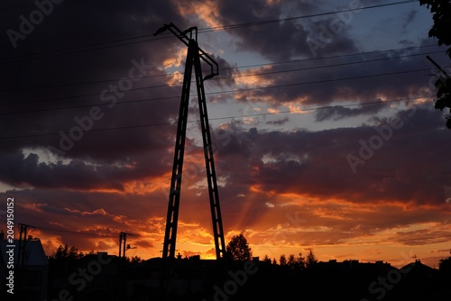 Power lines silhouette against dramatic sunset sky with dark clouds and fiery orange sunbeams over suburban houses