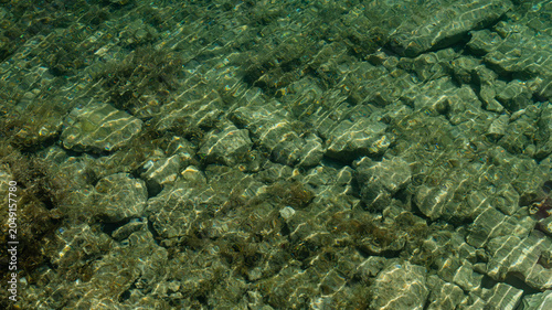 Top-down macro texture of crystal clear turquoise sea water with ripples and sun reflections over stones, Croatia.