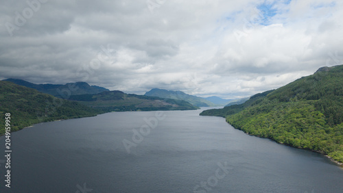 Photos Breathtaking aerial panorama of Loch Lomond from Firkin Point, showcasing the vast lake surrounded by majestic mountains and lush green forests under a dramatic cloudy sky in Scotland