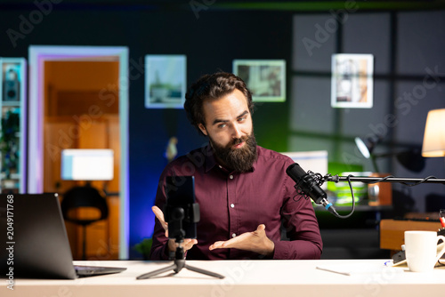 Media star sitting at table talking in front on cellphone on tripod stabilizer in dimly lit home studio interior  chatting with viewers. Influencer using phone to record vlog in living room