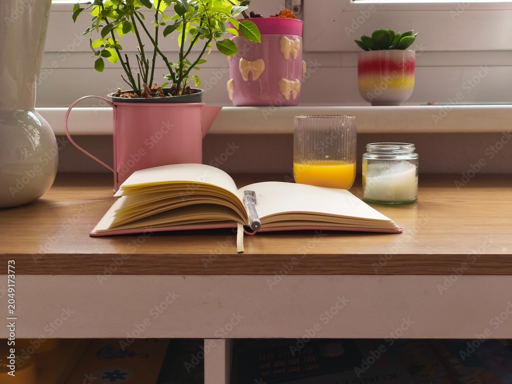 Fototapeta premium Open notebook with blank pages on wooden table beside potted plants, glass of orange juice, and candle in a cozy indoor setting